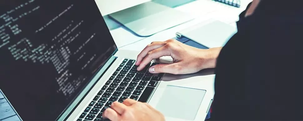Person typing code on a laptop with a monitor showing code.