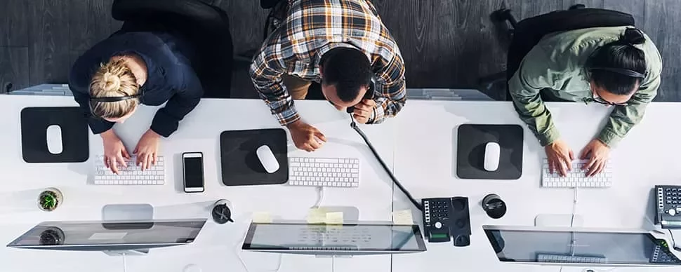 Overhead view of three people working at desks with computers and phones.