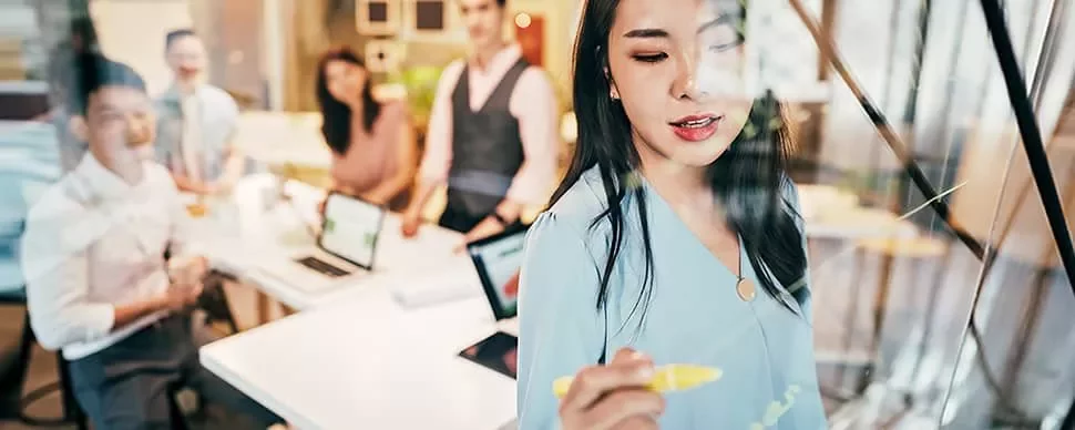Woman writing on glass board with a marker during a team meeting.