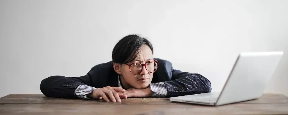 Man in glasses staring thoughtfully at a laptop on a wooden table.