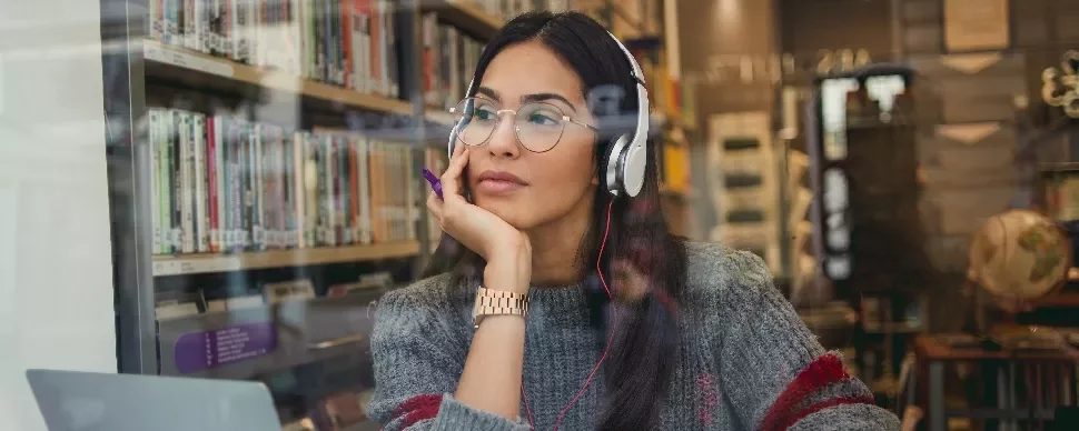 Woman with glasses and headphones, sitting in a library, looking thoughtful.