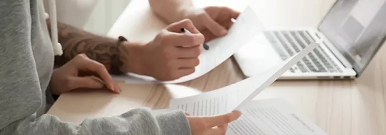 Two people reviewing documents at a desk with a laptop.