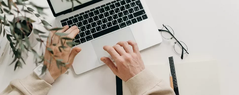 Top view of hands typing on a laptop, with glasses and a notebook on a white desk.