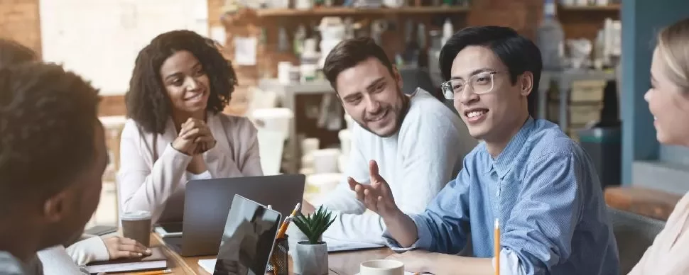 Group of people smiling and discussing at a meeting table with laptops and coffee cups.