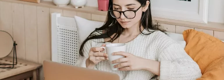 Woman wearing glasses, holding a mug, and using a laptop.