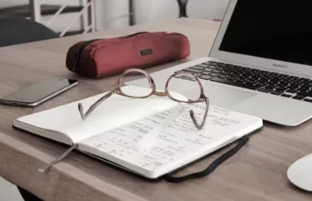 Notebook with eyeglasses on a desk next to a laptop and pencil case.